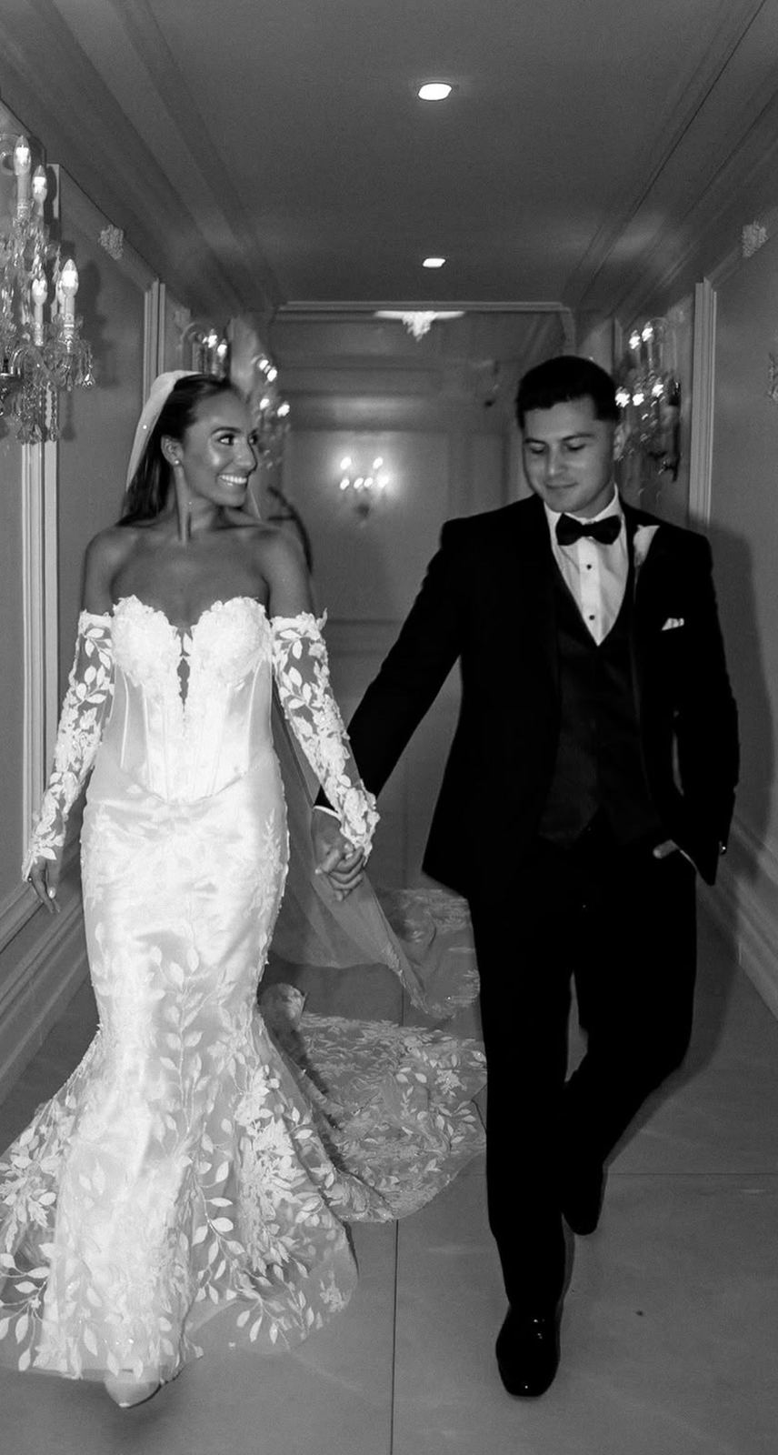 Pete and Cassie walking hand in hand down a chandelier-lit hallway, in black and white