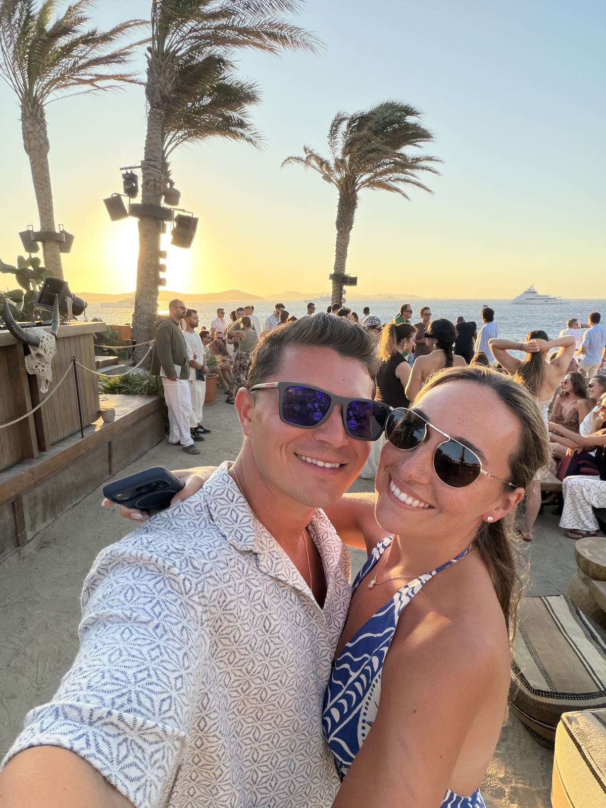 Pete and Cassie in sunglasses taking a sunset selfie at a beach club lined with palm trees